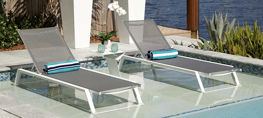 Sleek poolside lounge chairs with gray mesh fabric and blue striped towels, partially submerged in water, by a calm waterfront. The setting is enhanced by tropical plants, stylish planters, and a small side table with decorative items, creating a relaxing outdoor ambiance.