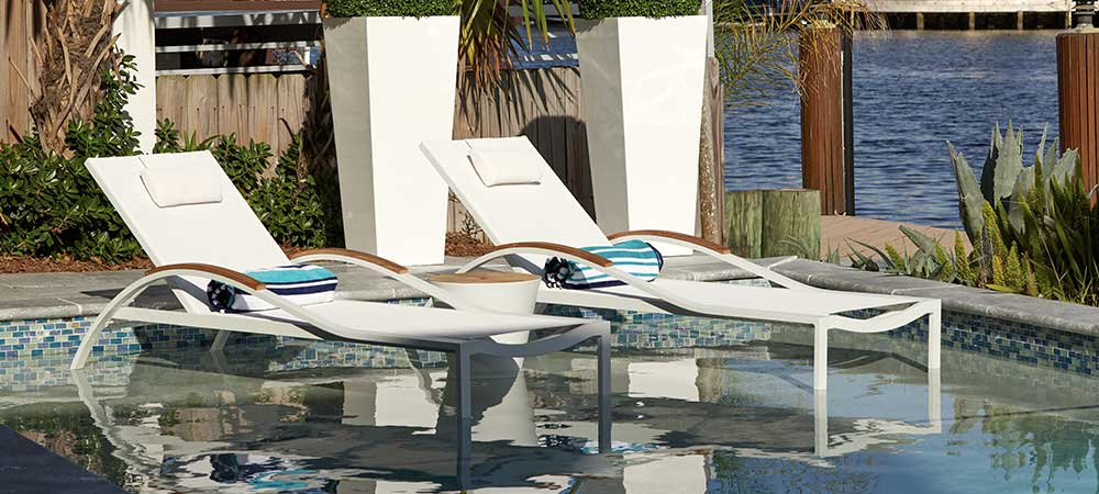 Modern poolside lounge chairs with white cushions and striped pillows, partially submerged in water, next to a serene waterfront. Surrounding tropical plants and stylish planters add to the tranquil outdoor setting