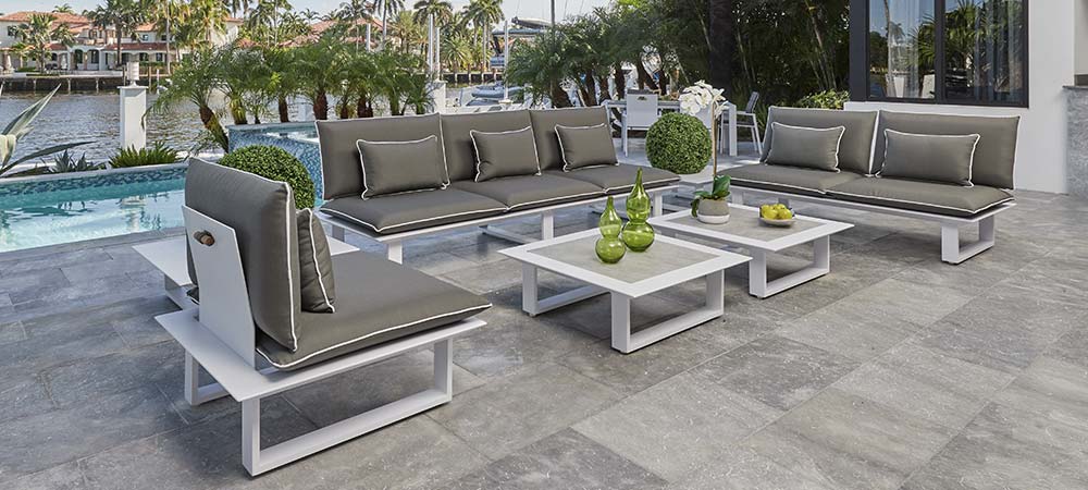 Outdoor patio area featuring modern white and gray sectional sofas with plush cushions, paired with matching coffee tables. The setting overlooks a beautiful waterfront with palm trees and a swimming pool, creating a luxurious and relaxing environment