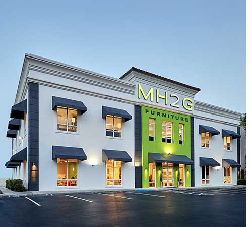 Exterior view of MH2G Furniture store in Fort Lauderdale, featuring a modern white building with dark gray accents, large windows with black awnings, and a bright green entrance sign.