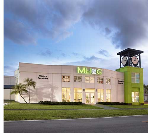 Exterior view of MH2G Furniture store in Miami, featuring a modern white building with large windows, a bright green entrance sign, and a distinctive clock tower. The surrounding area includes a palm tree and well-manicured lawns under a cloudy sky.