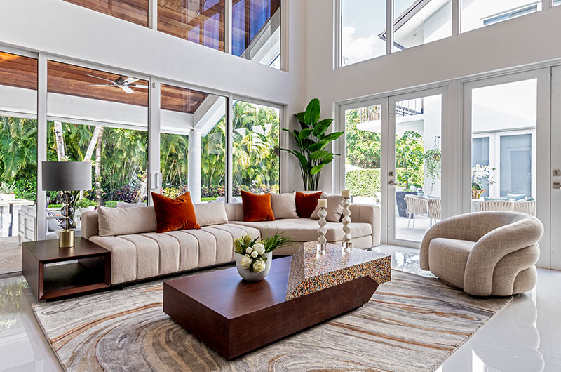 Modern living room with large windows, beige sofa, orange pillows, and wood coffee table.